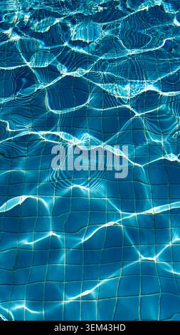 Clear blue swimming pool water surface with sunlight reflections creating ripples and patterns over white tiled floor beneath Stock Photo