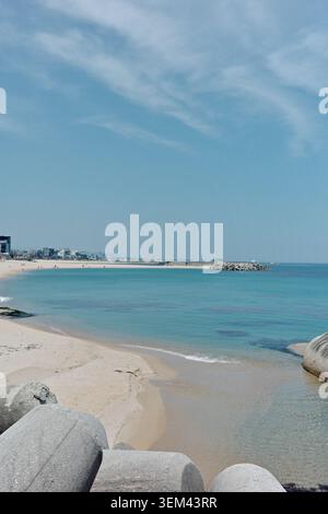 Wide sandy beach with turquoise sea, concrete tetrapod breakwater, and coastal town skyline under a hazy blue sky, East Sea, South Korea. Stock Photo