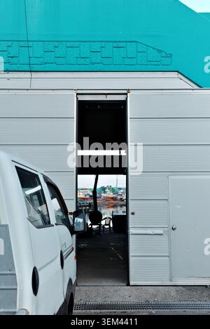White van parked beside a turquoise warehouse with open shutter door revealing fishing boats moored in a harbor, Gangneung, South Korea. Stock Photo