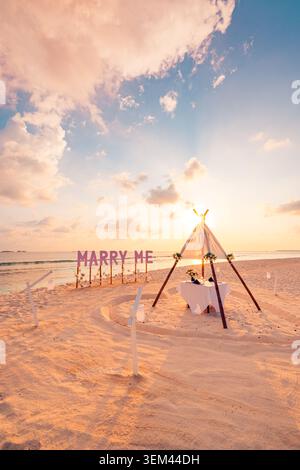 Romantic marry me sign beach wedding proposal setup. Tropical sunset palm trees teepee decoration. Engagement honeymoon getaway celebration scene Stock Photo