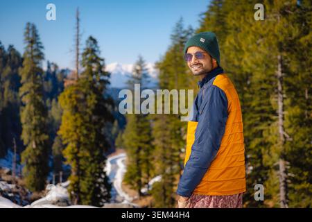 A happy man wearing a warm jacket stands in a snowy pine forest during a bright winter day in Kashmir, India. Stock Photo