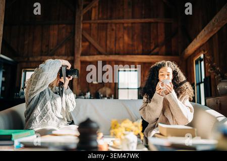 Woman photographing friend enjoying coffee in cozy cabin Stock Photo