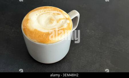 Close-up view of a white ceramic coffee cup filled with frothy cappuccino on a dark textured surface Stock Photo