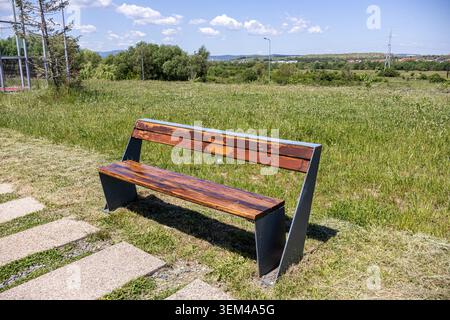 An outdoor horizontal view of a stylish new park bench made of dark polished wooden planks and a charcoal metal frame, positioned next to a modern sto Stock Photo