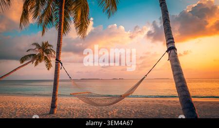 Hammock hanging between palm trees at golden sunset beach, dramatic evening clouds over calm sea, peaceful tropical island relaxation scene scenic Stock Photo