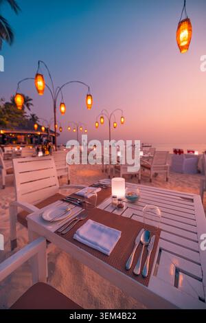 Hanging lanterns above beach dinner table at dusk vertical view, elegant place settings on sandy shore, warm romantic tropical resort evening scenic Stock Photo