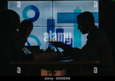 Business teammates analyzing data and collaborating on strategy in a dim meeting room with charts Stock Photo