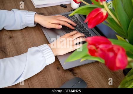 Close side view of female hands typing on laptop keyboard near red tulips on wooden desk. Online education, remote work, productivity and modern digit Stock Photo