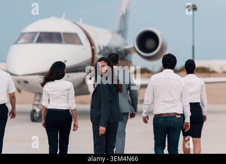 Joyful businesswoman in dark suit glancing back over shoulder while diverse flight crew and executives walk toward private jet across sunlit airport Stock Photo