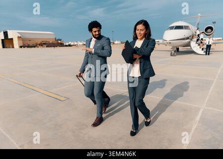 Engaged business colleagues a man and woman in tailored suits reviewing notes on a tablet folio while striding away from a private jet across a sunlit Stock Photo
