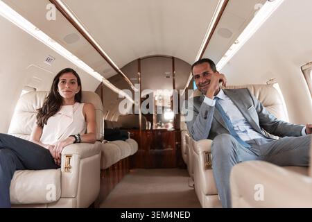 Professional multiethnic business woman in white sleeveless top and man in grey suit sitting comfortably in beige leather seats of luxury private jet Stock Photo