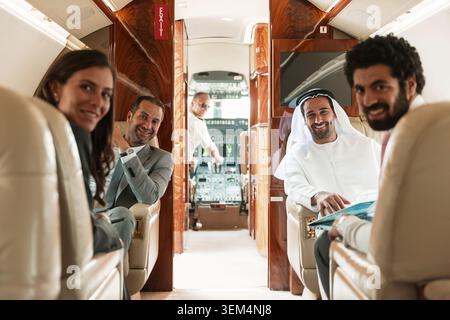 Diverse multiethnic business travelers including traditional Emirati partner smiling and working on laptop inside luxurious private jet cabin with Stock Photo