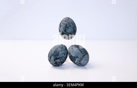 Three blue marbled Easter eggs styled as cosmic or stone pattern on white background. Close-up of naturally dyed textured eggs with one egg on a small Stock Photo