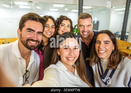 Six coworkers are posing for a selfie in a bright and modern coworking space. The atmosphere is friendly and relaxed, with visible workspace elements Stock Photo