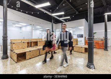 In a contemporary coworking space, multiple individuals walk past wooden seating areas while engaging in their activities. Stock Photo