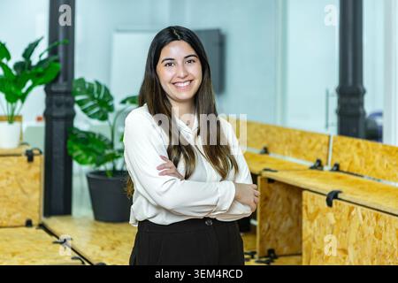 A young woman stands confidently in a coworking space. She smiles, arms crossed, while surrounded by wooden desks and plants. Stock Photo