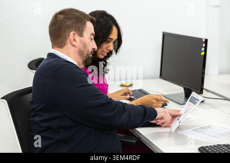 Two coworkers are engaged in discussion while reviewing documents at a shared workspace. The setting is designed for collaboration, featuring computer Stock Photo