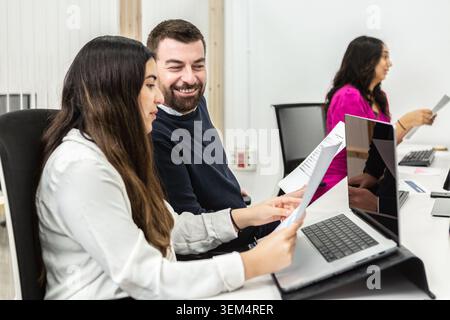Two professionals work together in a coworking space, focused on reviewing documents. They sit at desks equipped with laptops and papers in a bright, Stock Photo