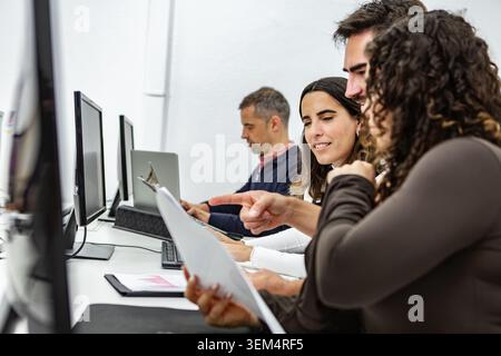 In a coworking space, four individuals are seated at desks, working on their computers. They are engaged in a discussion, pointing at documents and sh Stock Photo