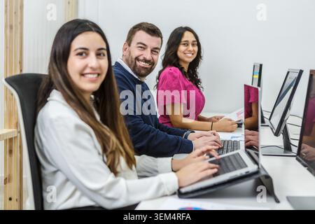 Three coworkers sit at modern desks in a well-lit coworking space. They are focused on their laptops while engaging in discussions. Stock Photo