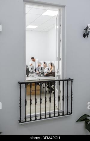 A group of four people works in a bright, minimalist coworking space. They are focused on their computers, collaborating on projects. Stock Photo