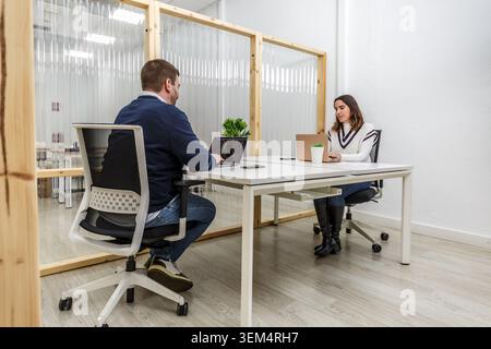 Two individuals work in a coworking space featuring a modern design with wooden partitions. One person uses a laptop while the other focuses on a tabl Stock Photo