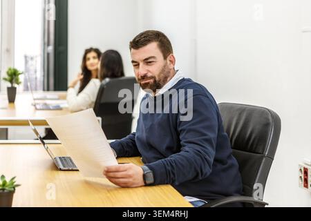 A man looks over documents in a coworking space. Two women are seated in the background, working on their laptops. Stock Photo