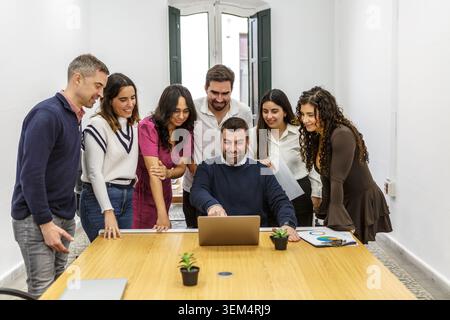 A group of seven coworkers gather around a table in a modern coworking space. They are engaged in a discussion and working together on a laptop. Stock Photo