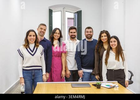 Seven coworkers stand together in a modern coworking space. They smile and engage with each other, showcasing a collaborative atmosphere. Stock Photo