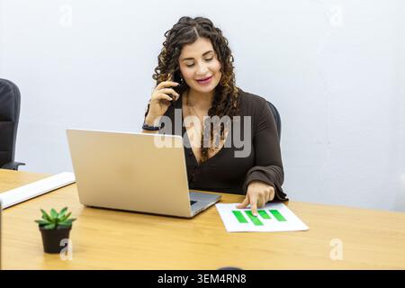 A young businesswoman is seated at a wooden desk in a coworking space. She is speaking on the phone and reviewing financial charts printed on paper. Stock Photo
