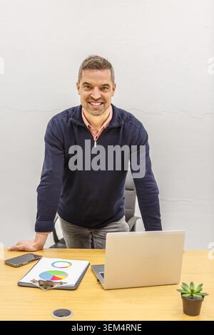 A man is working in a coworking space, standing near a desk featuring a laptop, documents, and a plant. The workspace offers a modern environment for Stock Photo