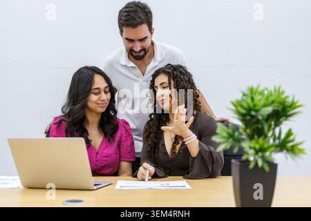 Three professionals are engaged in a collaborative work session in a coworking space. They are seated at a wooden table with laptops and documents. It Stock Photo