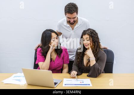 Three individuals are engaged in a discussion at a coworking space. They are reviewing documents and analyzing data on a laptop. Stock Photo