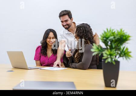 Three professionals are gathered around a table in a coworking space. They are reviewing documents on a laptop, discussing a project. Stock Photo