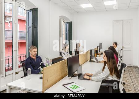 A group of six professionals are engaged in work at a coworking space during daylight hours. The office features modern desks and large windows. Stock Photo