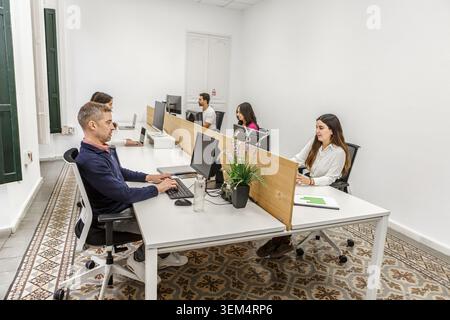 A group of five people works in a modern coworking space. They are focused on their computers, seated at shared desks. Stock Photo