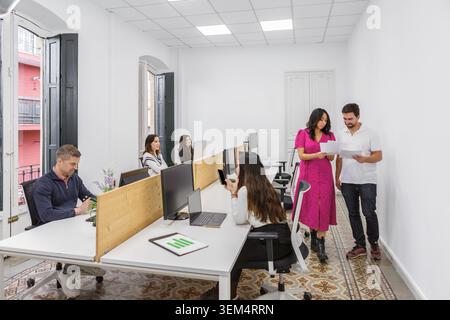 A group of six professionals are engaged in various activities within a modern coworking space. It is daytime in a well-lit room with desks, computers Stock Photo