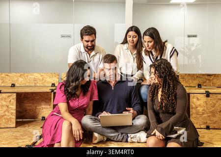 A group of six professionals engages in collaboration at a coworking space. They are gathered around a laptop on a wooden floor while discussing ideas Stock Photo