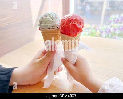 Two hands holding red and green ice cream cones together beside a window Stock Photo