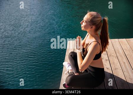 A woman is sitting cross-legged on a wooden dock, practicing yoga by the water. She enjoys the warm sunlight. Stock Photo