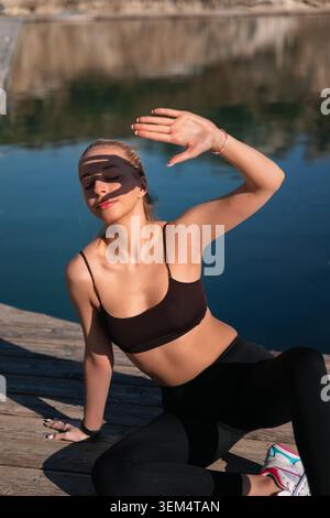 A woman wearing sports clothes is seated by the water in the afternoon light while exercising and enjoying the sun. Stock Photo