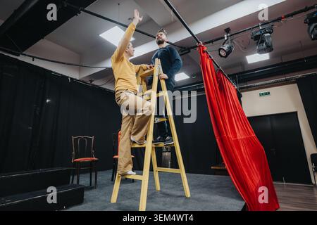 Young actors rehearsing on stage as one climbs a ladder beside a red curtain Stock Photo