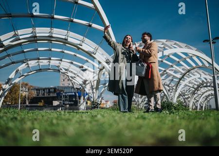Two work colleagues enjoying coffee and taking a selfie outdoors under modern architectural arches Stock Photo