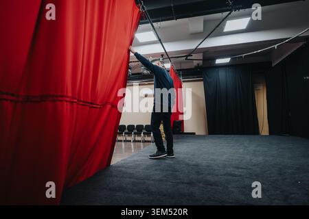 Young actors opening red curtain on empty theatre stage during rehearsal Stock Photo