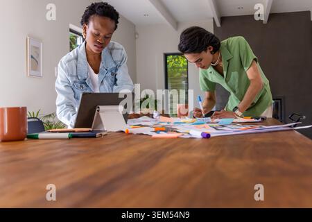 Diverse coworkers leaning over table, referencing tablet on stand and writing sticky notes at home. Collaboration, planning, brainstorming, workspace, Stock Photo
