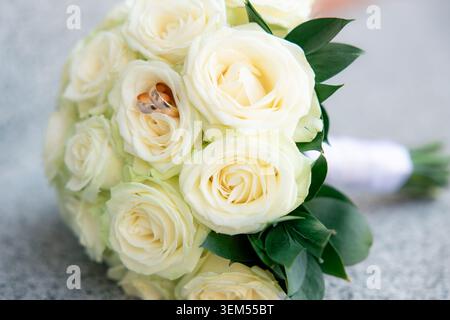 A bridal bouquet of white roses and greenery cradles a pair of wedding rings in soft natural light Stock Photo