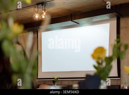 Indoor wedding or event venue with projection screen on the wall, rustic timber ceiling beams and industrial Edison bulb pendant lights. Yellow flower bokeh in the foreground adds a romantic, atmospheric touch to this darkened event space. Stock Photo