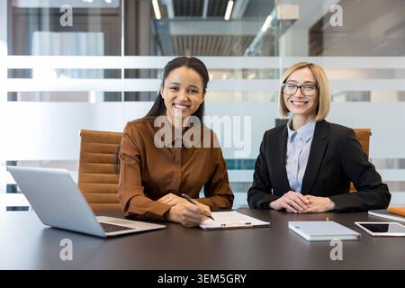 Two cheerful businesswomen at a bright office table, one signing a document while the other smiles at the camera, conveying teamwork, confidence and professional collaboration Stock Photo
