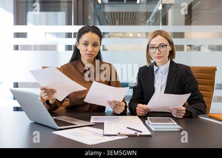 Two diverse businesswomen reviewing documents and looking at the camera during a corporate meeting, representing professionalism, teamwork, and project management in a contemporary workspace Stock Photo