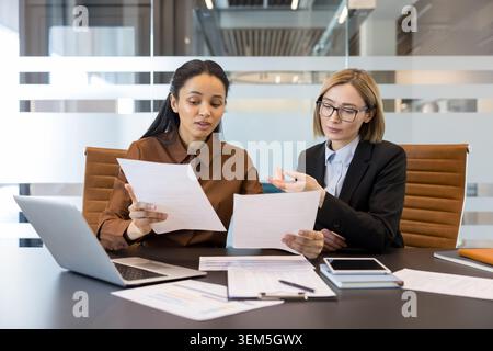 Two professional businesswomen collaborating and analyzing financial documents during a corporate meeting in a contemporary office setting, focusing on teamwork and strategy Stock Photo